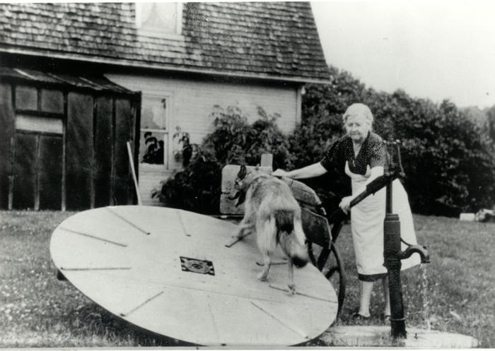 Black and white image of dog on circular platform pumping water. Older woman stands behind.