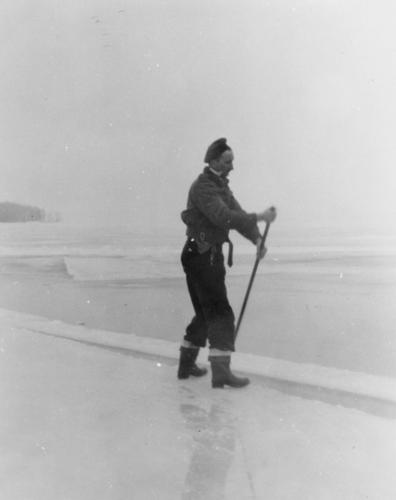 Photo of Wallace Wright in rubber boots and a cap cutting ice on Lake Simcoe and floating ice blocks down the channel to the elevator using a long pole.