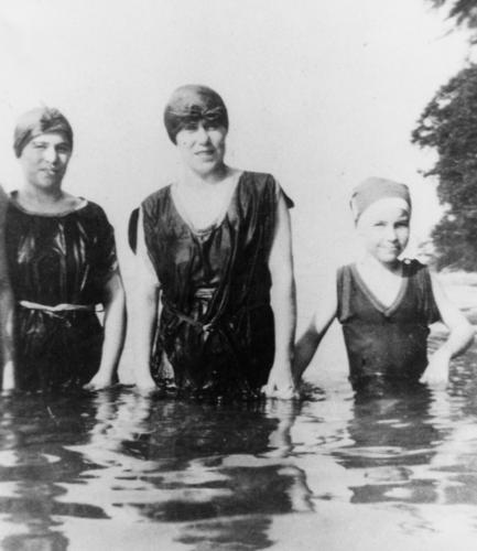 2 women and a girl in old fashioned bathing suits and bathing caps standing waist deep in a lake