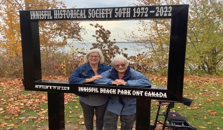 Two women stand behind a life-sized photo frame at Innisfil Beach Park recognizing the 50th Anniversary of the Innisfil Historical Society