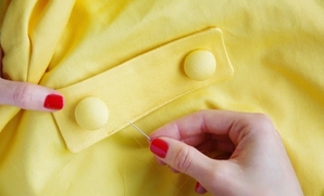 Image of a woman's hand with red fingernails sewing a button onto a yellow textile