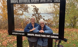 Two women stand behind a life-sized photo frame at Innisfil Beach Park recognizing the 50th Anniversary of the Innisfil Historical Society